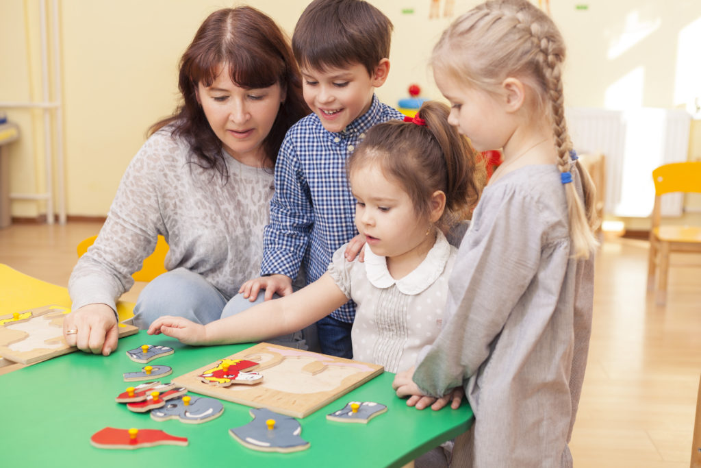 beautiful mature woman teacher collect puzzle with three pupils of a kindergarten classroom at the green table. Horizontal color image.